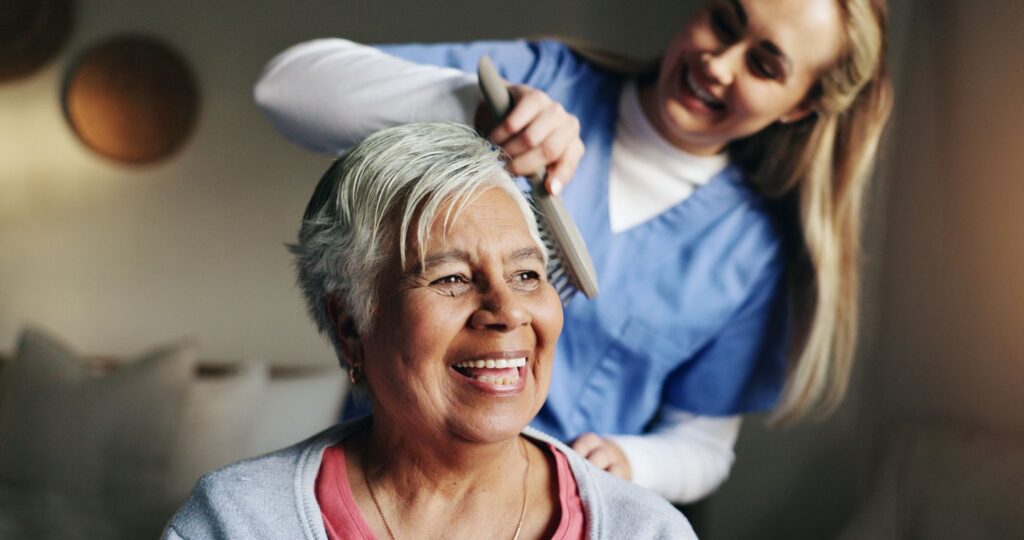 A senior woman smiles as an in-home caregiver brushes her hair.